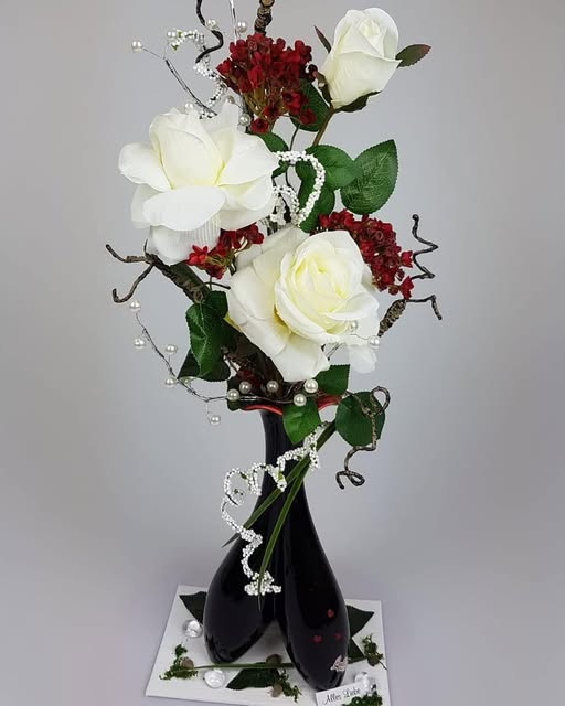 Weiße Rosen, grüne Blätter und kleine rote Akzente in einer hohen schwarzen Vase auf einer weißen dekorierten Unterlage.