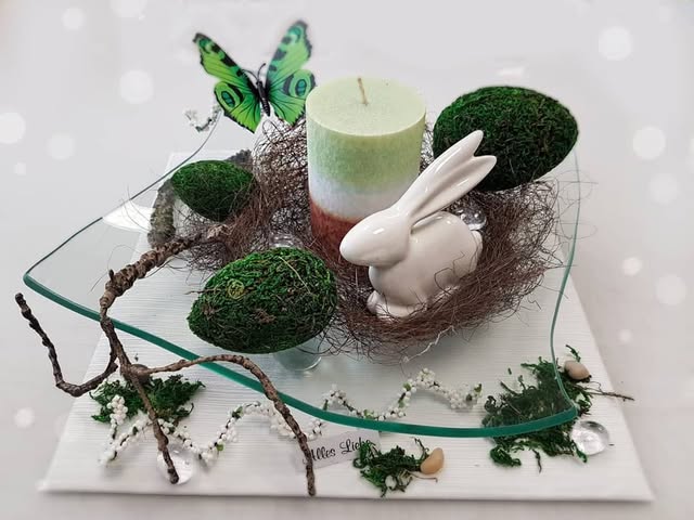 Frühlingsarrangement auf einer Glasplatte mit weißem Hasen, hellgrüner Kerze, grünen Kugeln, Zweigen und Schmetterling.